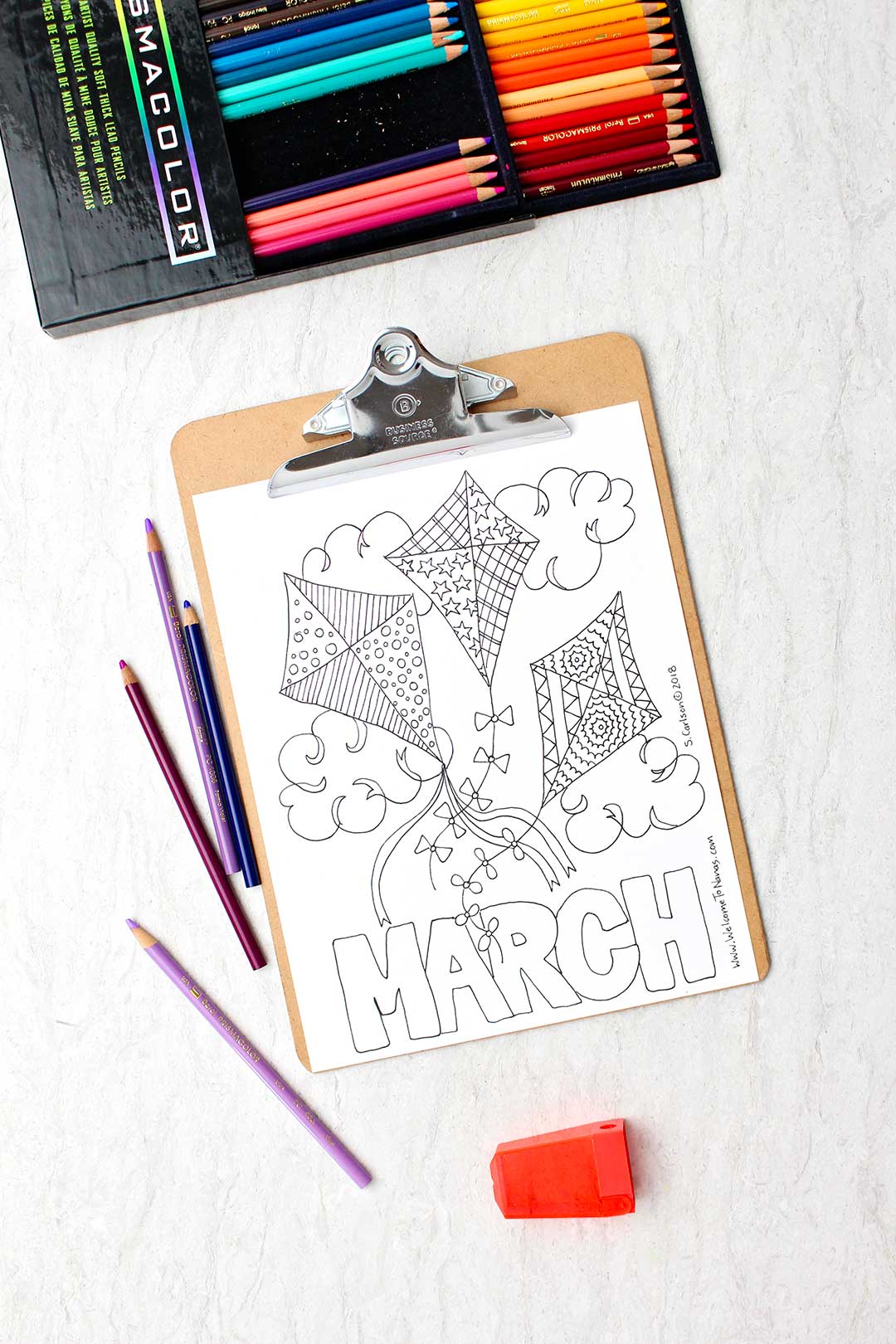 An uncolored March coloring page on a clipboard with colored pencils nearby. The page shows three patterned kites in the sky with the word "March" at the bottom.