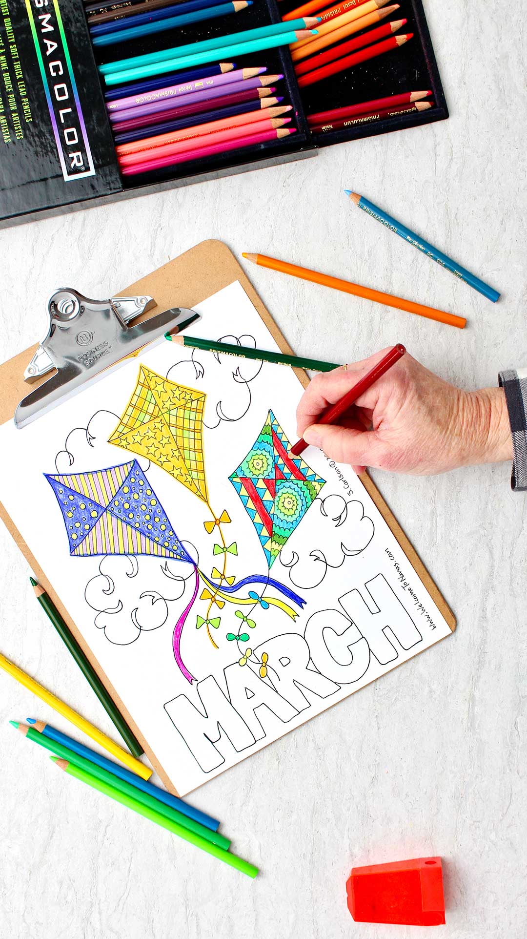 Child coloring a design on the kite on the right side using a red colored pencil. All three kites are colored in.