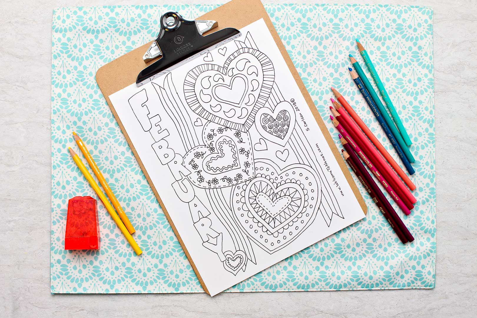 Uncolored February Coloring Page on a clipboard with colored pencils close by.