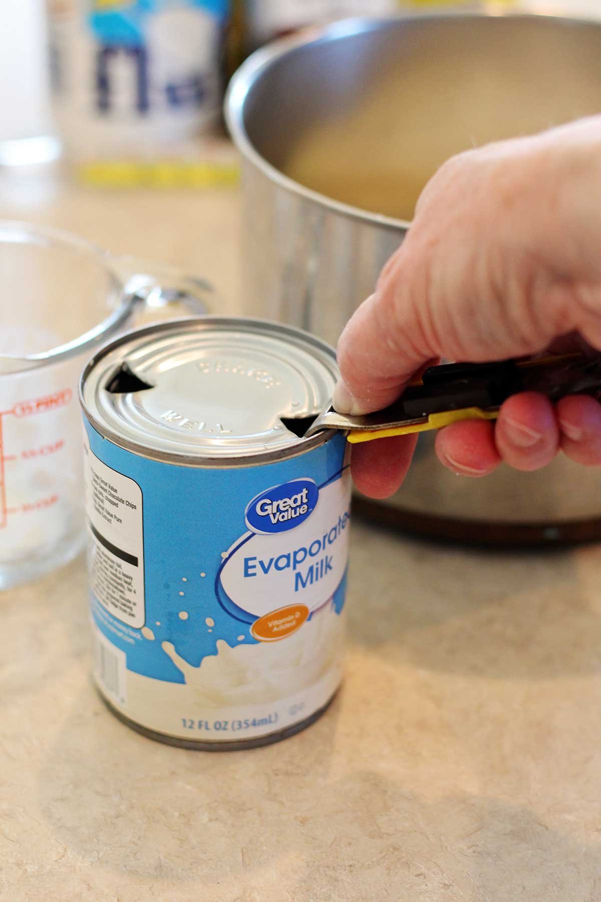A person using a can opener on a can of evaporated milk with a pot in the background.