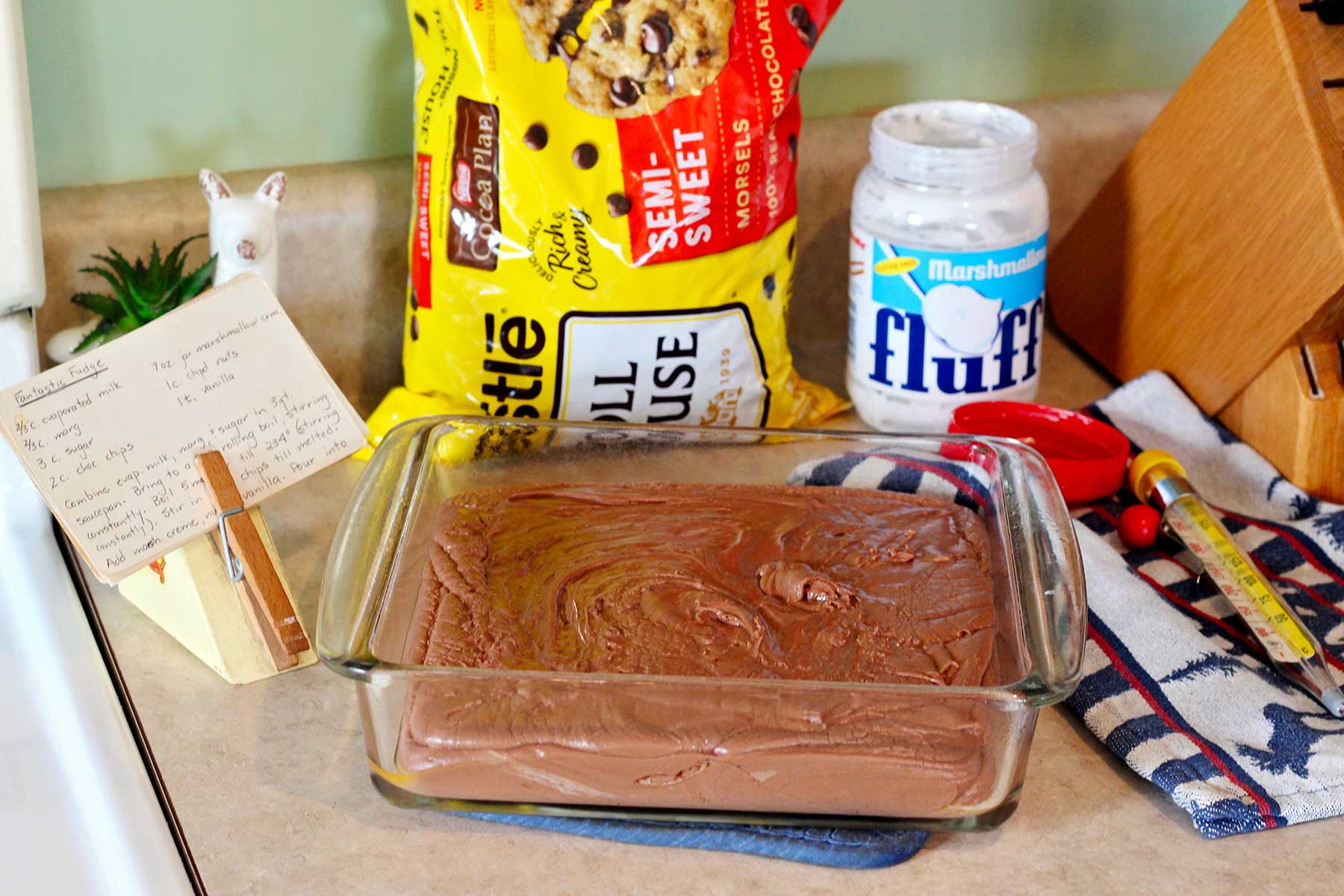 Fudge mixture in glass baking pan on counter with chocolate chips, fluff and recipe near by.
