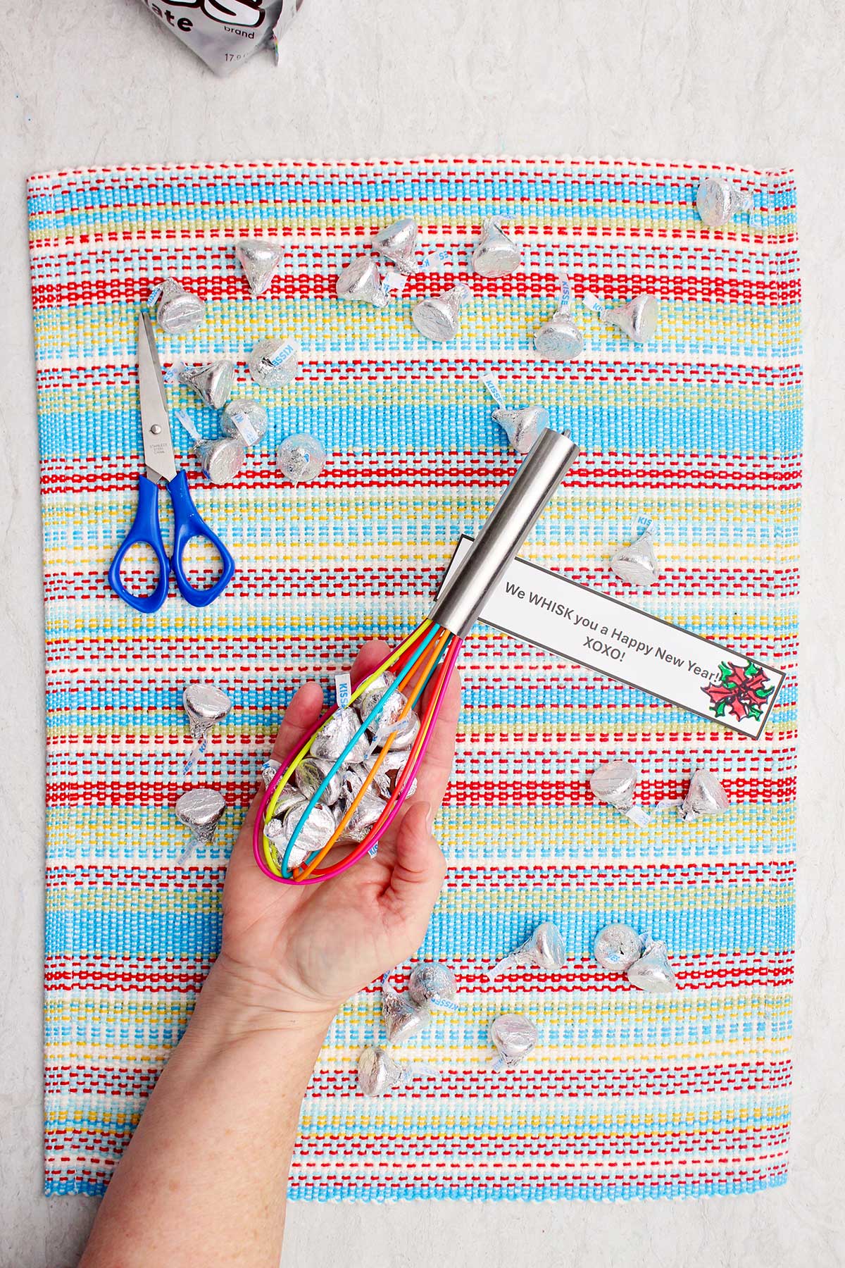 A person holding a whisk full of Hershey Kisses with other candies scattered on a colorful striped place mat.