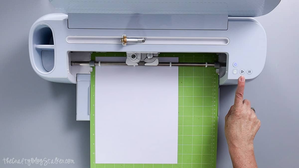 Cricut cutting machine slicing white cardstock on a green cutting mat while a hand engages the start button.