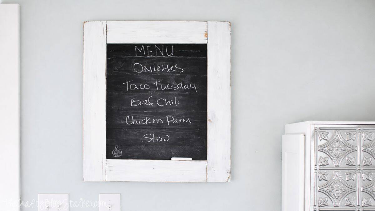 Farmhouse chalkboard menu with a white wood frame listing dishes like omelettes, taco Tuesday, beef chili, chicken parm, and stew.