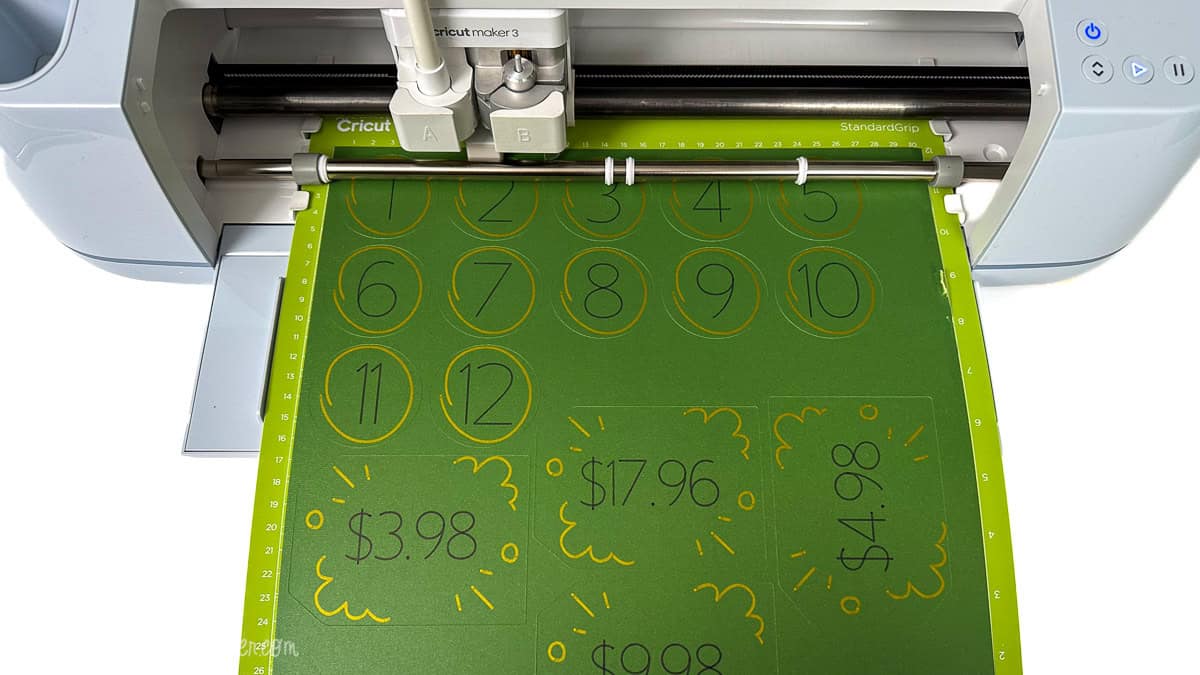 Cricut Maker 3 cutting machine sketching numbered circles and price tag designs on cardstock using a Cricut pen for a DIY Price Is Right baby shower game.
