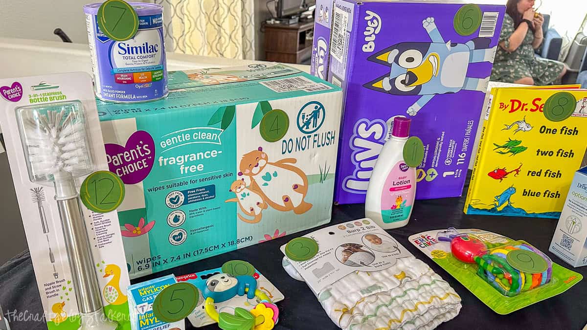 Baby shower game arrangement with baby products labeled by number, including diapers, wipes, baby lotion, formula, book, toys, and bottle brush for a Price Is Right guessing game.