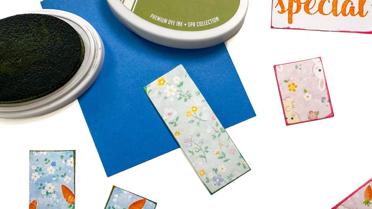 Ink pads and small patterned paper pieces on a table, indicating edges being inked before assembling a handmade card.