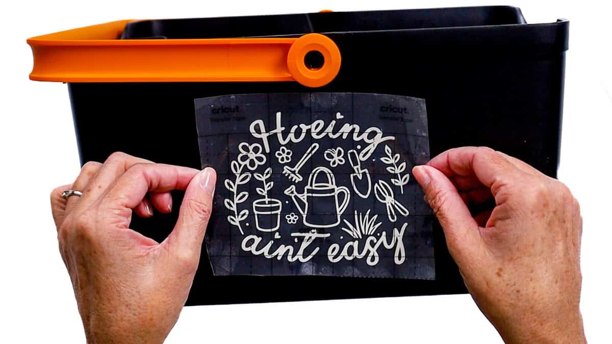 Hands position a gardening-themed vinyl decal with transfer tape onto the front of a black garden bucket before pressing it into place.