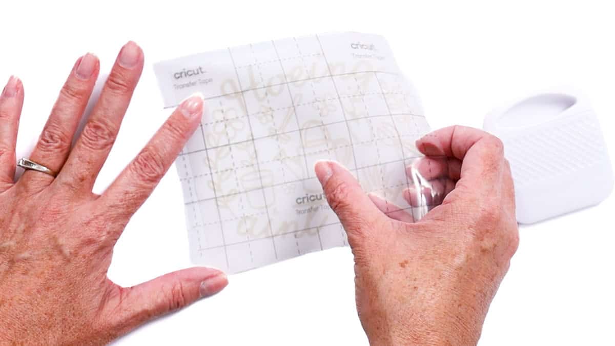 Hands apply transfer tape over a weeded vinyl decal, smoothing it down with a scraper tool in preparation for application.