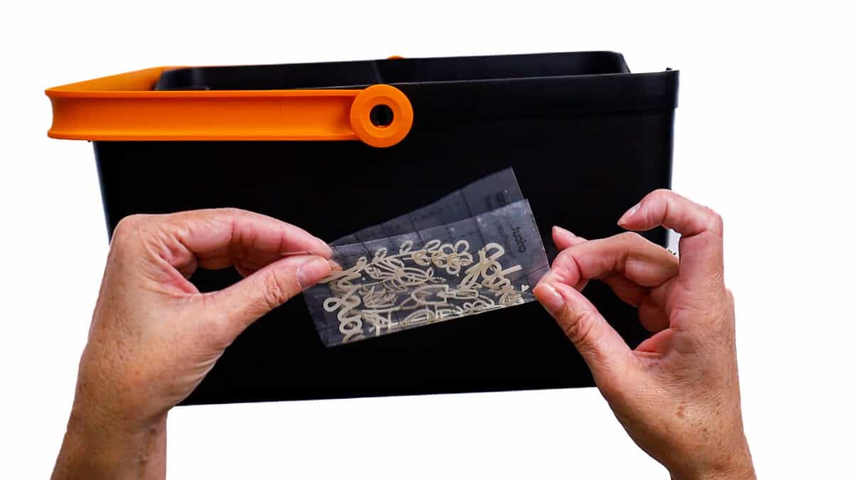 Hands hold a small vinyl decal with transfer tape in front of a black garden bucket, aligning it to find the center before applying.
