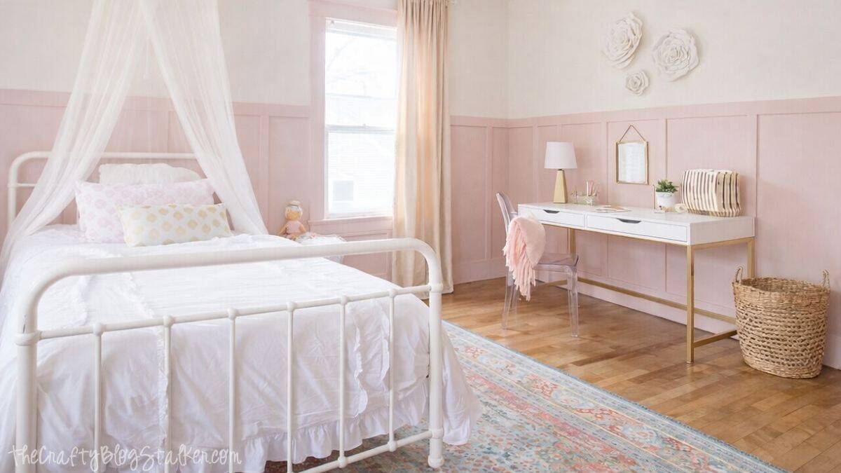Styled girls bedroom featuring a white metal bed with a canopy, window drapes, a desk with chair, wall decorations, a woven basket, and a patterned rug on timber floors.