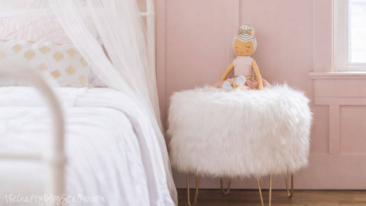 Stuffed toy resting on a fluffy upholstered stool beside a white metal bed with sheer canopy drapes in a softly styled girl's bedroom.