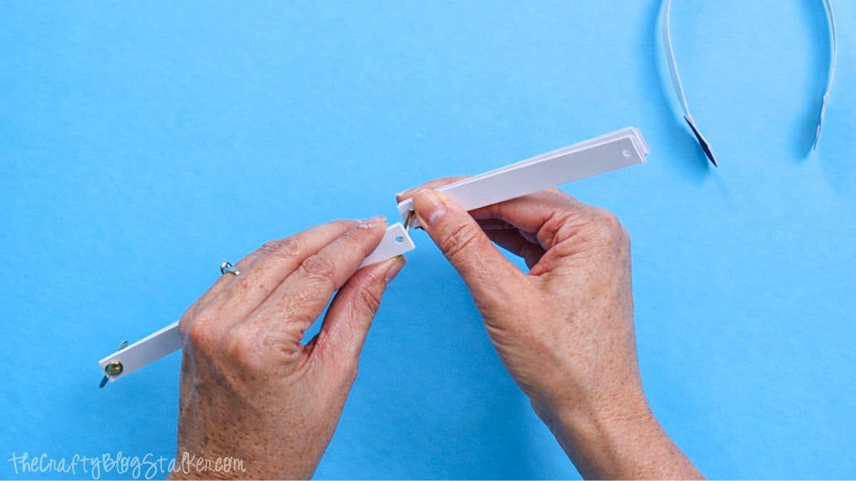 Hands linking two white paper strips with a metal brad fastener, forming a connection for a paper strip snowman craft on a blue work area.