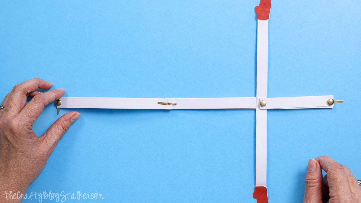 Hands organizing white paper strips joined with metal brads in a cross formation, with mitten cutouts on the ends, creating the snowman arm and body framework.