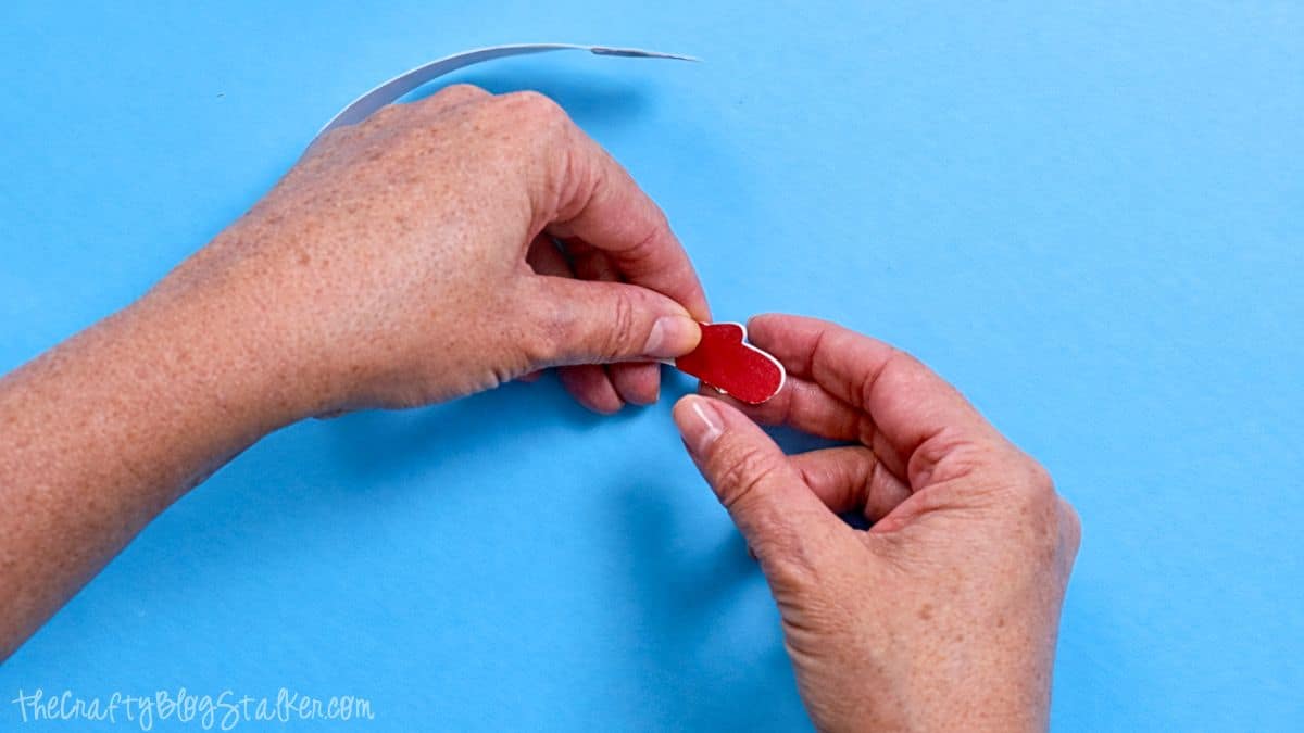 Hands attaching a small paper mitten to a curved paper strip, assembling components of a paper strip snowman craft on a crafting surface.