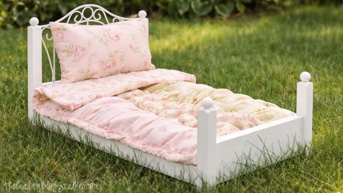Handcrafted ruffled quilt and cushion on a small white doll bed situated on grass, illustrating a completed doll bedding collection outdoors.