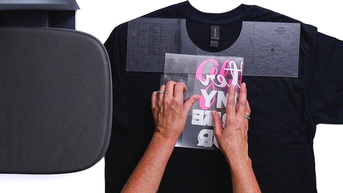 Hands arranging an iron-on vinyl design on a T-shirt utilizing alignment guides, with a heat press platen close by prior to pressing.