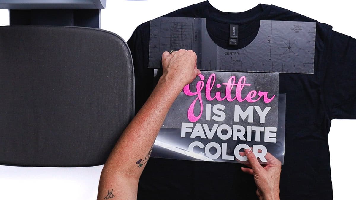 Hands centering an iron-on vinyl design on a T-shirt using a T-shirt alignment ruler, with a heat press platen nearby the shirt.