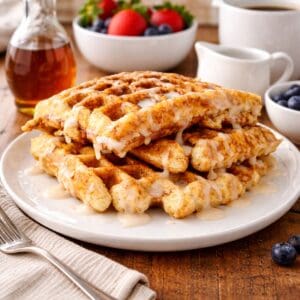 Tower of cinnamon swirl waffles drizzled with icing on a plate, paired with syrup, fresh berries, and coffee on a wooden surface.
