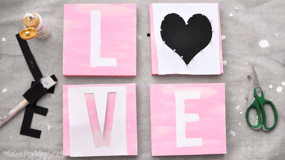 Four painted wooden blocks arranged to form LOVE, accompanied by letter stencils, a heart template, paint, brush, tape, and scissors during a DIY banner project.