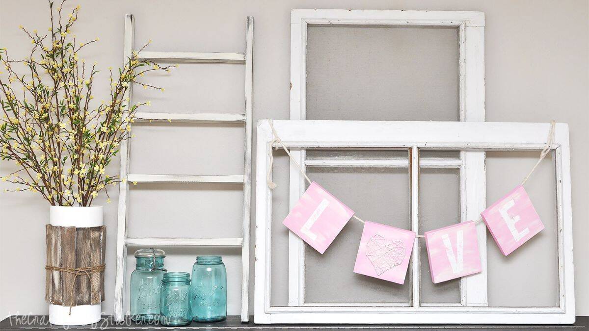 Valentine’s mantel decoration featuring a DIY LOVE banner displayed on a vintage window frame, surrounded by a rustic ladder, glass jars, and a floral arrangement.