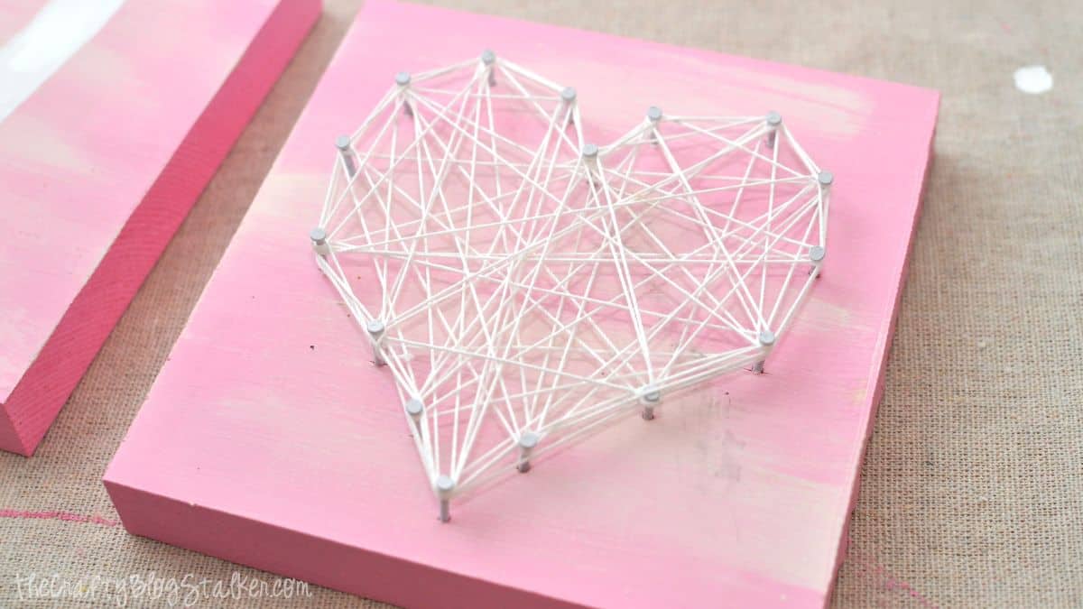 Heart-shaped string artwork created using nails and wrapped string on a painted wooden square, showcased as part of a DIY Valentine decor endeavor.