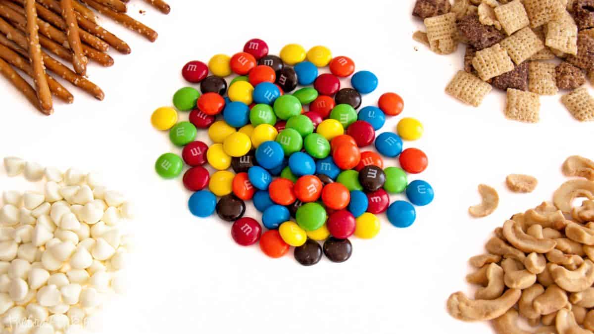 M&M White Chocolate Party Blend Recipe Components for white chocolate party mix placed on a white surface, including pretzels, cereal squares, white chocolate chips, cashews, and candy-coated chocolates.
