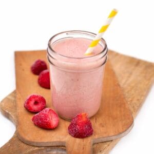 Strawberry smoothie in a mason jar with a striped paper straw, set on a wooden cutting board with frozen strawberries beside it.