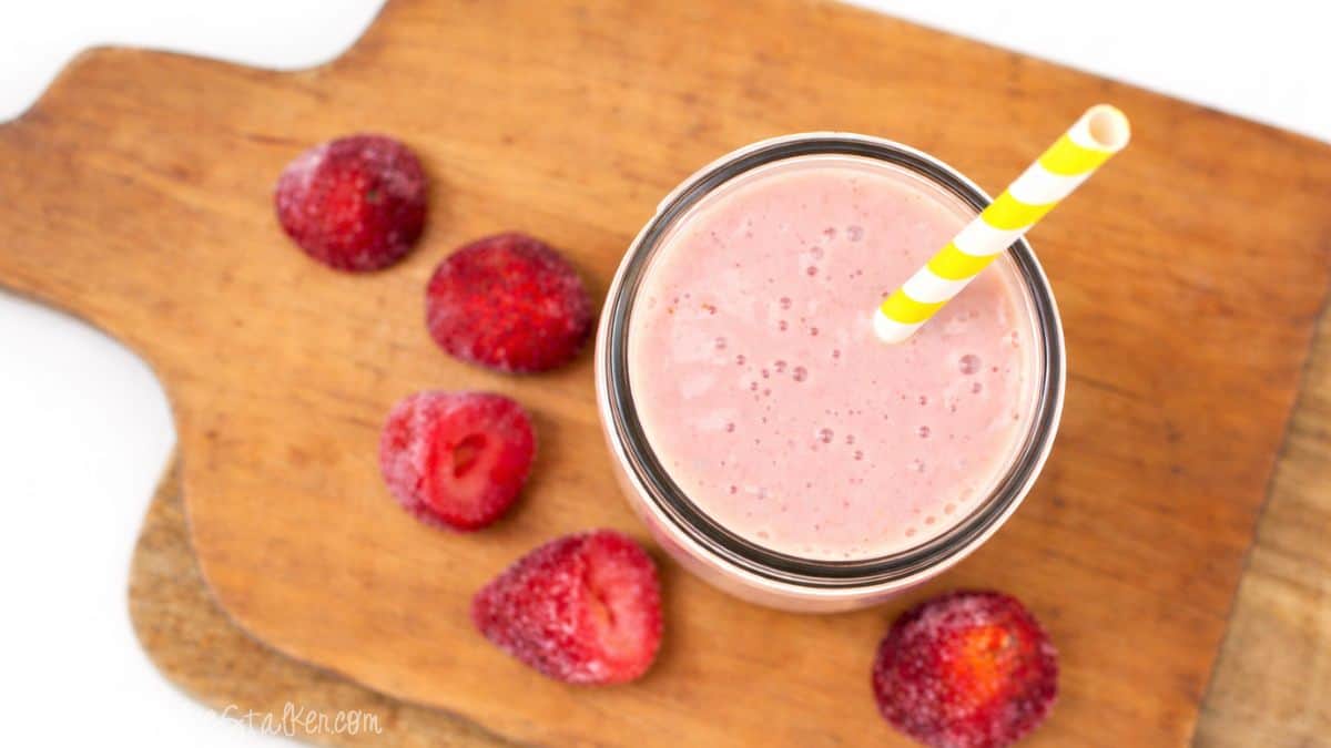 Aerial view of a strawberry smoothie in a mason jar with a striped paper straw, positioned on a wooden cutting board surrounded by frozen strawberries.