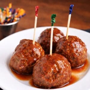 Sweet chili meatball snacks on a white plate, each adorned with a toothpick and blanketed in a sweet sauce, with further picks blurred in the backdrop.
