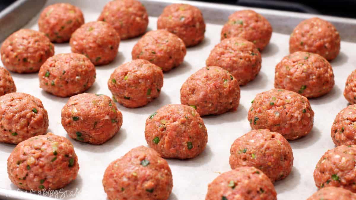 Uncooked meatballs organized on a parchment-lined baking tray, prepared for baking, featuring uniformly sized meatballs with herbs blended in.