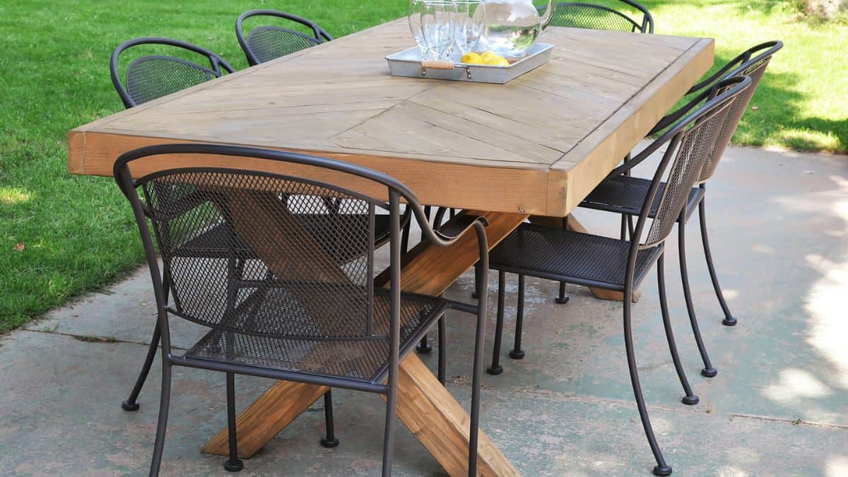 Crafted wooden outdoor dining table featuring a herringbone top and X-style base, surrounded by metal patio chairs on a concrete deck.