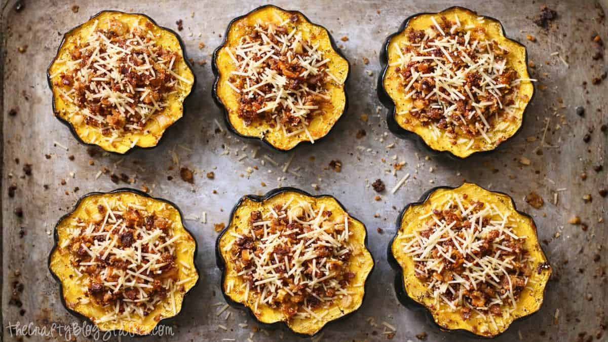Six halved acorn squashes filled with sausage stuffing and grated cheese, arranged on a baking tray ready for roasting.