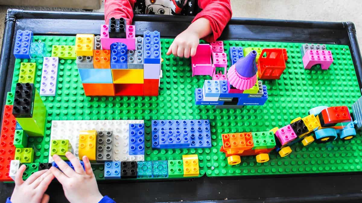 Children engaging with LEGO bricks on a DIY LEGO table, constructing colorful designs and vehicles on a large green baseplate.