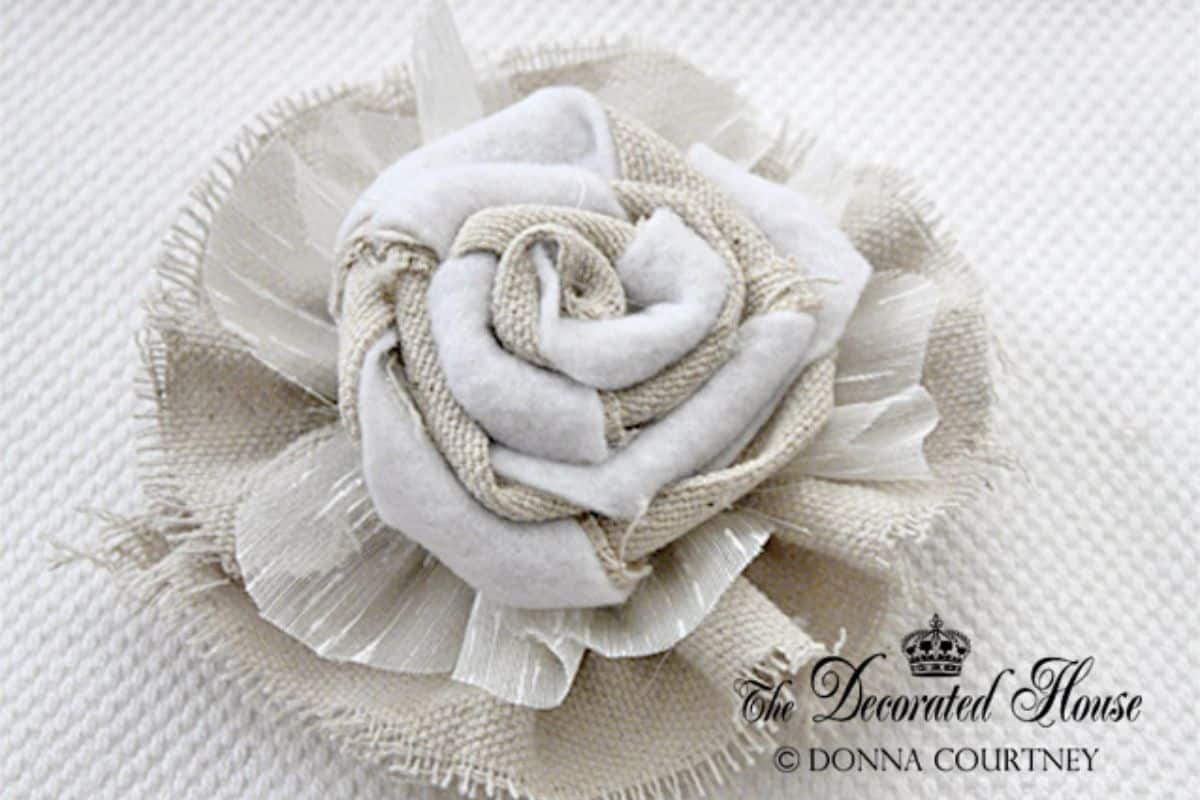 Artisan fabric flower composed of layered burlap, felt, and translucent material, styled in a rose formation against a textured backdrop.