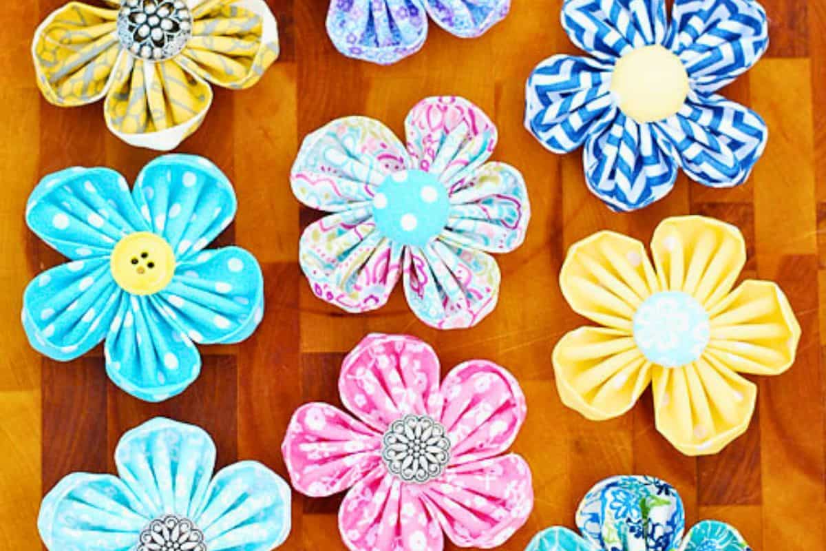 An assortment of handmade round petal kanzashi fabric flowers with button centers, artistically arranged in a flat lay on a wooden surface as a DIY craft illustration.
