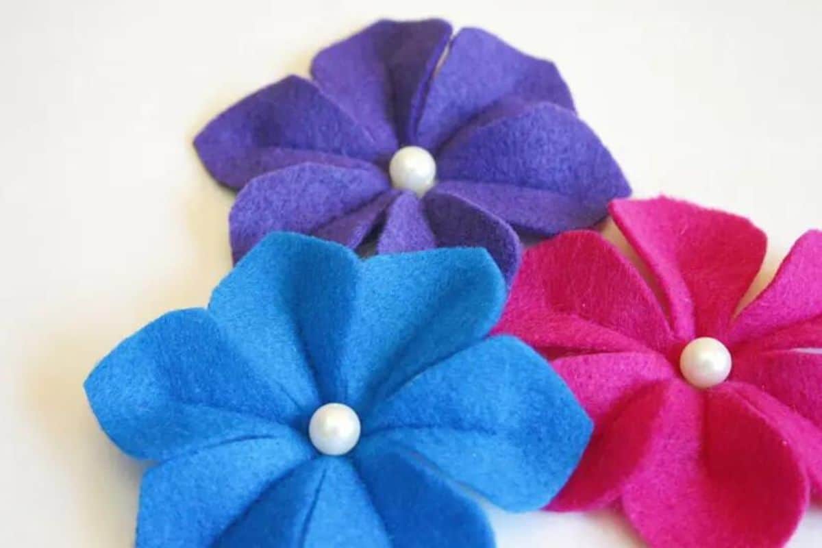 Three handcrafted felt camellia flowers featuring layered petals and pearl-like centers set against a light backdrop as DIY craft accents.