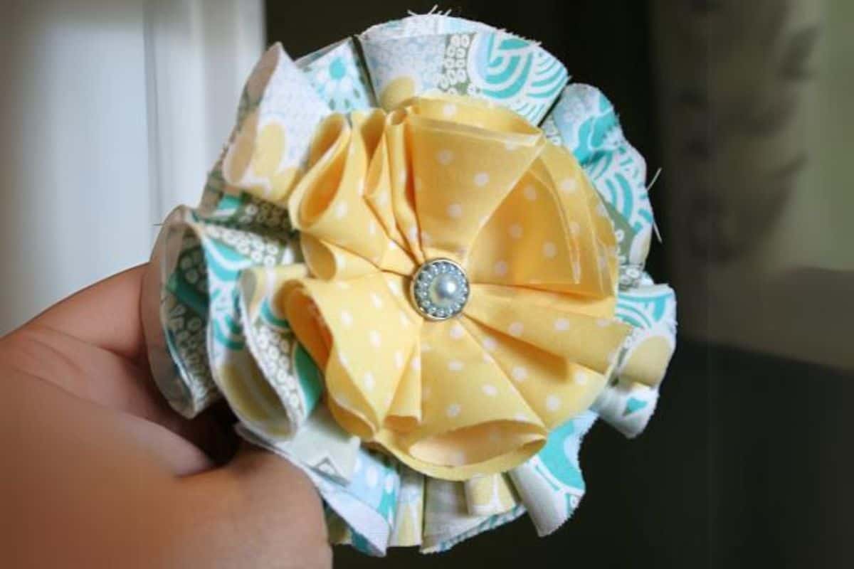 A hand presenting a layered fabric flower crafted from folded patterned cotton featuring a decorative button center, displayed as a close-up handmade craft.