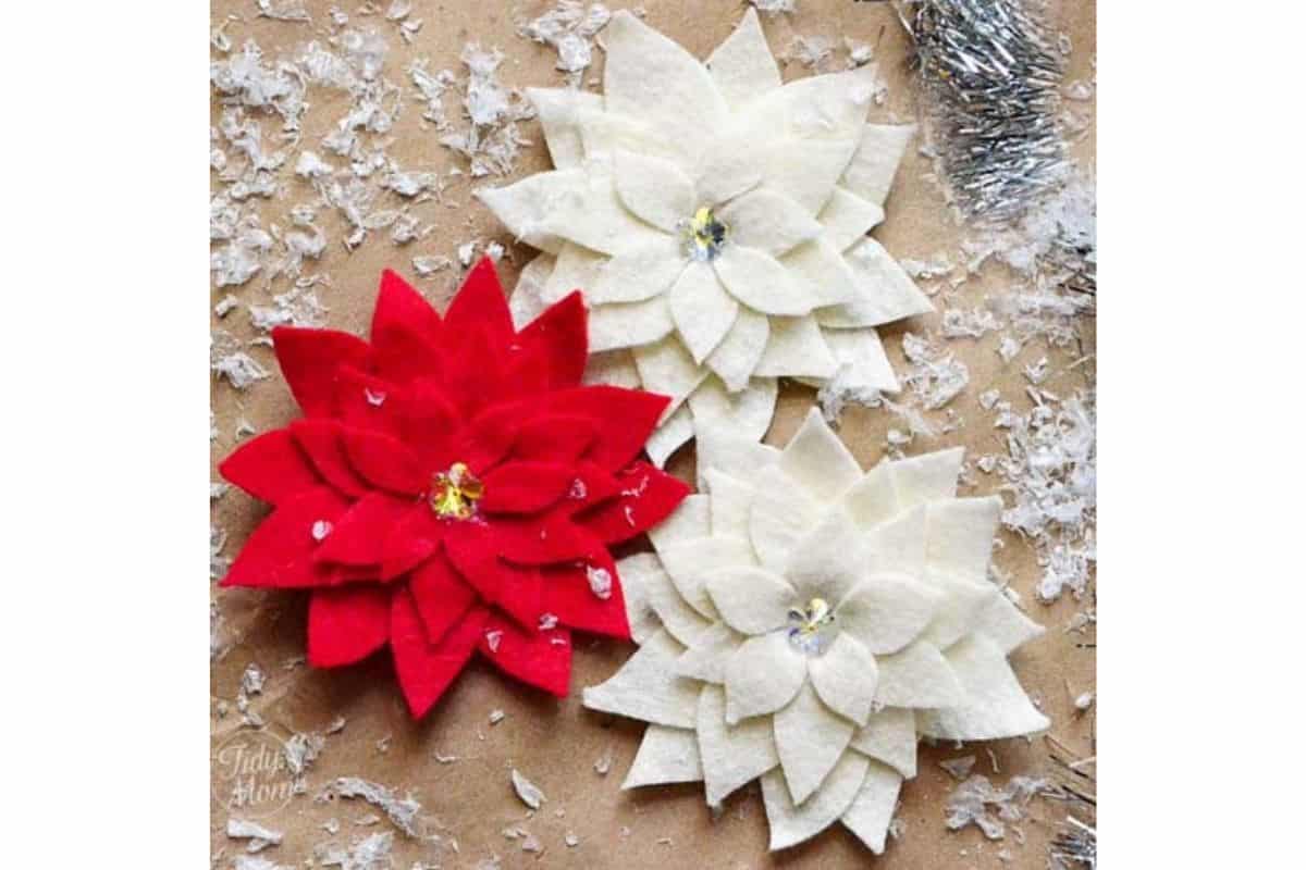 Handcrafted felt poinsettia blooms arranged on a rustic surface scattered with faux snow, displaying layered petals and decorative centers for festive decor or adornments.