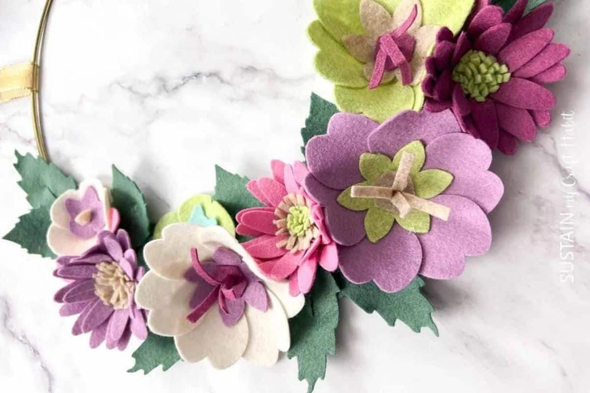 Golden hoop wreath adorned with handcrafted felt flowers and layered leaves, clustered together on a marble-like surface.