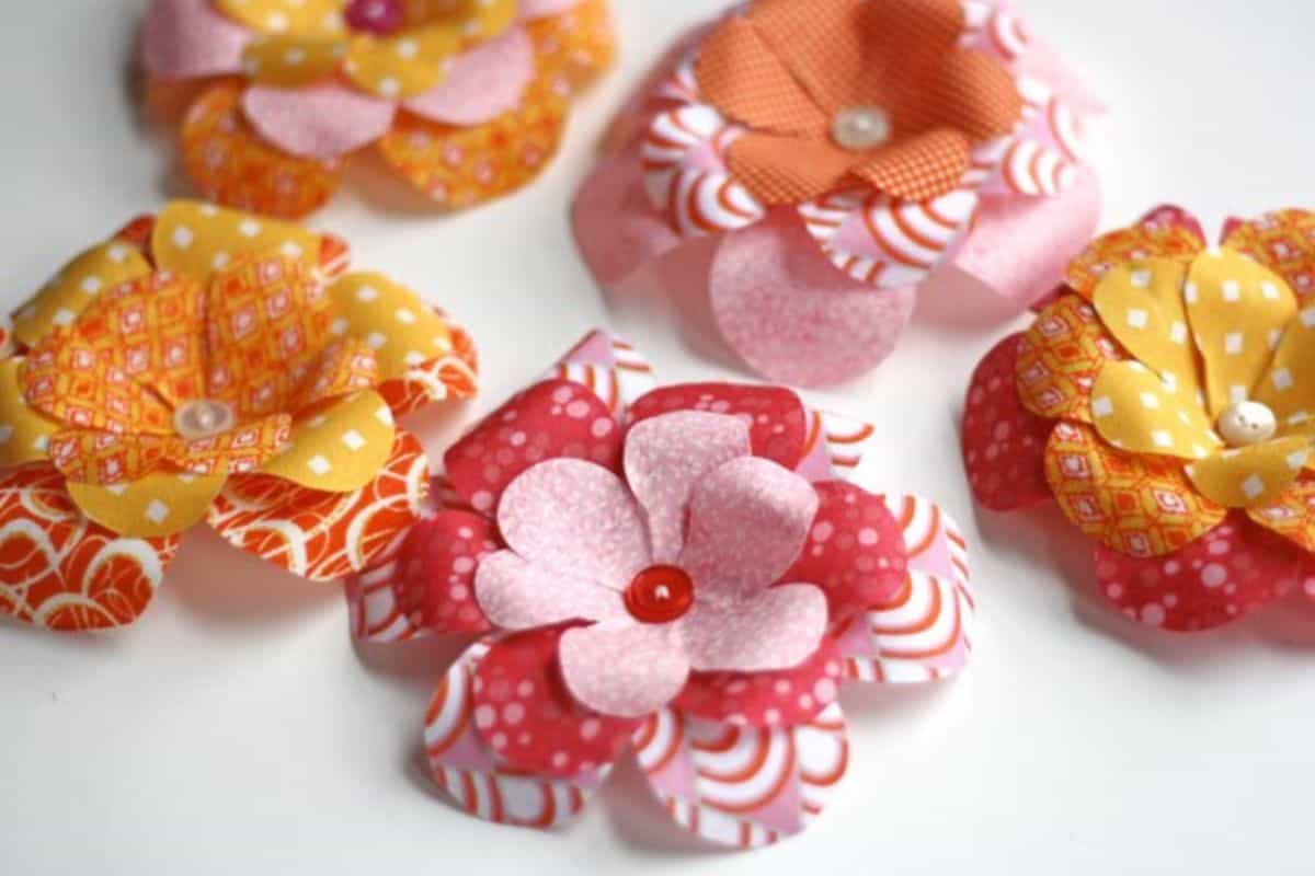 A collection of handmade fabric flowers featuring layered petals and buttons or bead centers, grouped together on a white surface as a craft exhibit.