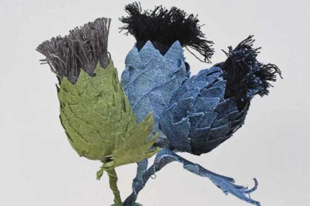 Three crafted fabric thistle flowers with textured foliage and fringe-like tops, gathered together against a plain backdrop.