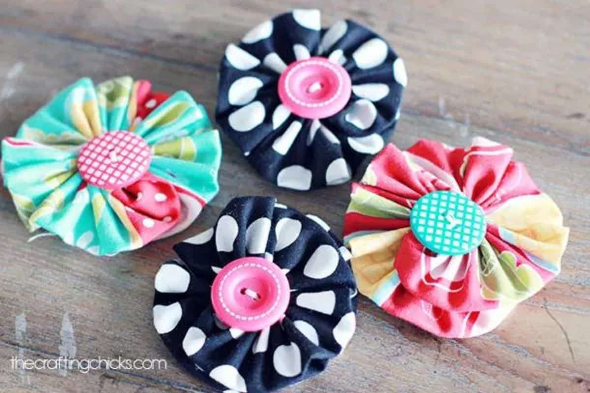 Four handcrafted yo-yo style fabric blooms featuring gathered circles and button centers arranged on a wooden tabletop as DIY craft accents.
