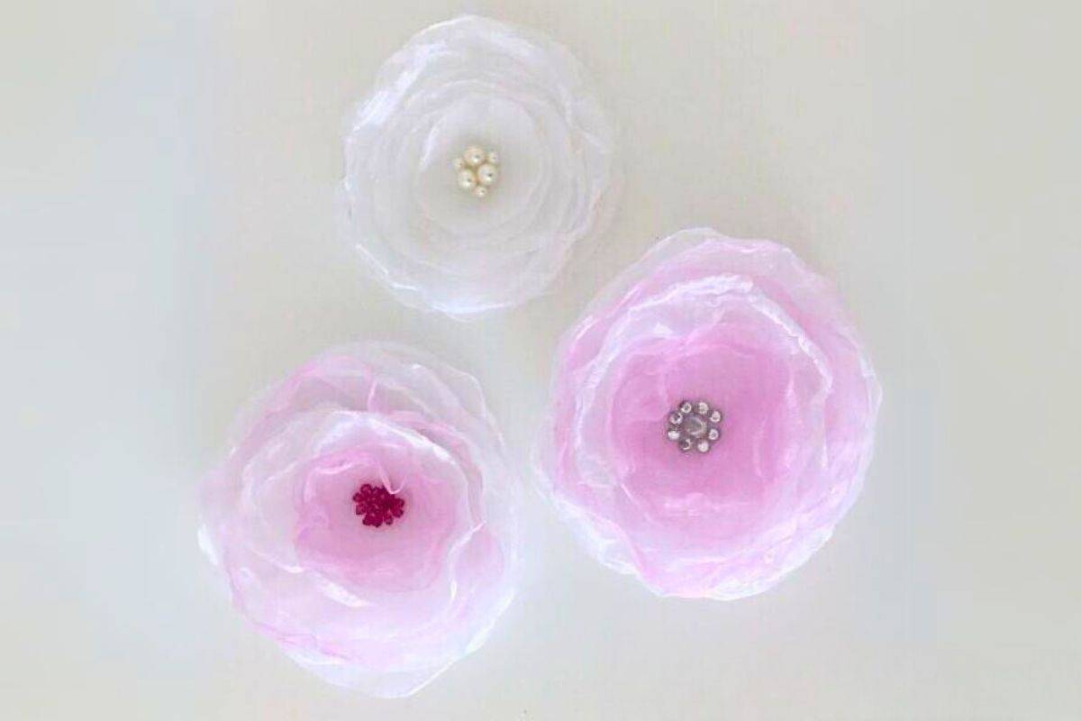 Three handmade organza fabric flowers displayed on a flat surface, each featuring layered sheer petals and bead centers, utilized as decorative accents.