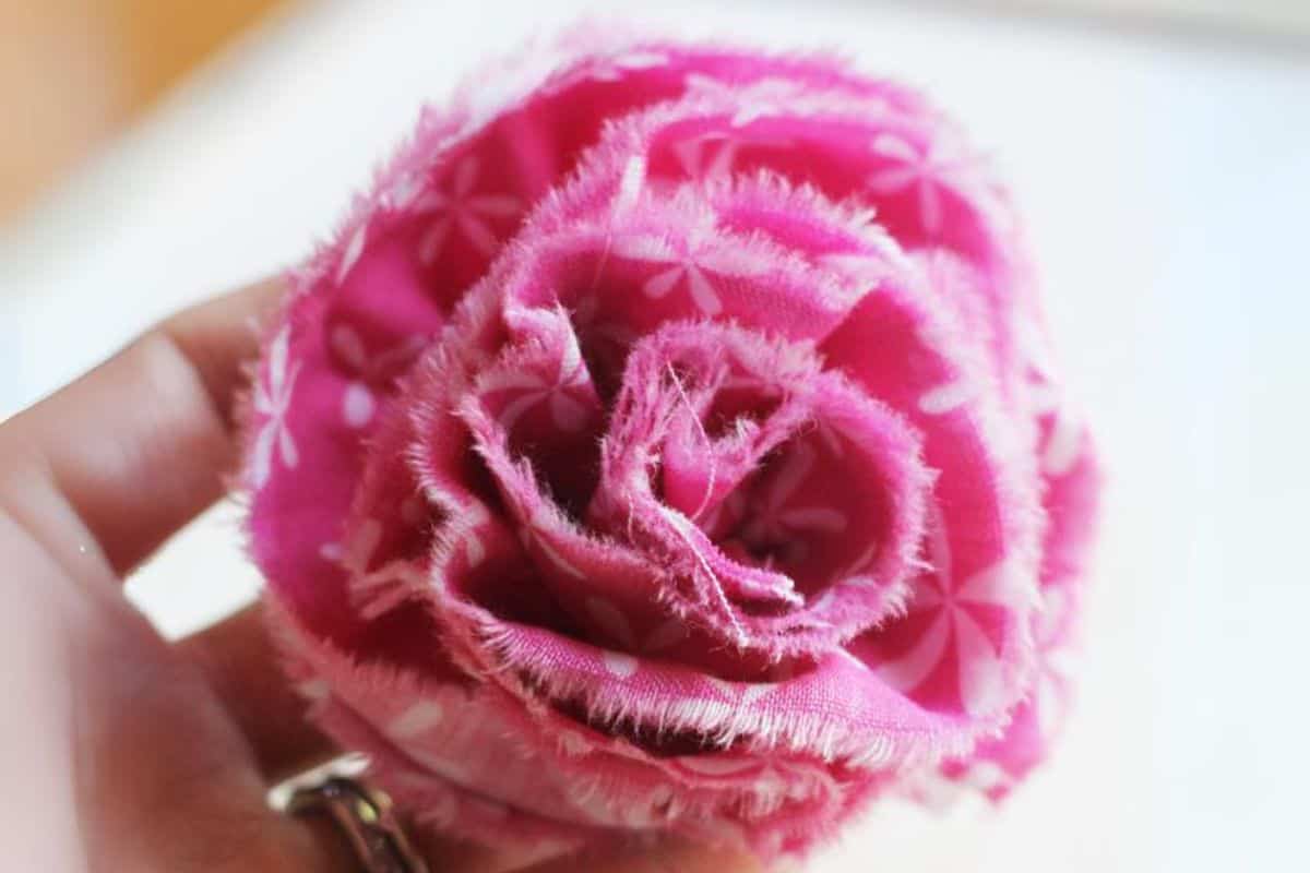 Close-up of a handcrafted ruffled fabric flower with frayed edges, held between fingers, illustrating layered petals and stitched details.