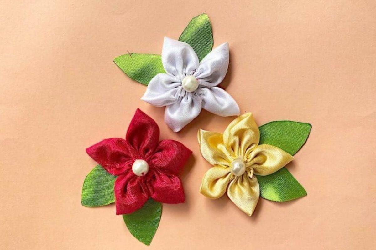 Three crafted satin fabric flowers with stacked petals and green leaves arranged on a solid surface, each adorned with a pearl-like center.