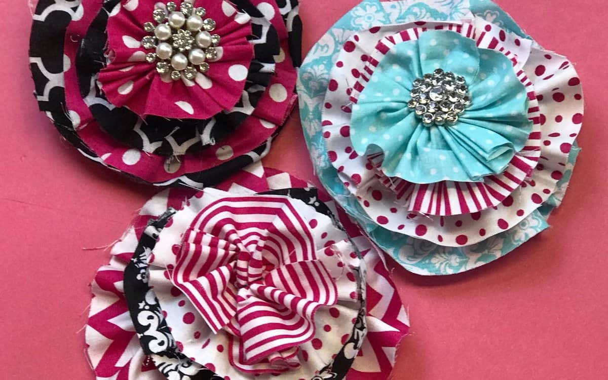 Three layered handmade fabric blooms with patterned petals and sparkling centers, displayed on a flat surface as a DIY craft illustration.