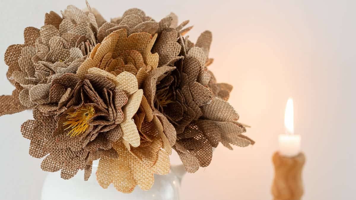 Bouquet of handmade burlap flowers arranged in a glass jar, featuring layered fabric petals and a softly blurred lit candle in the background.