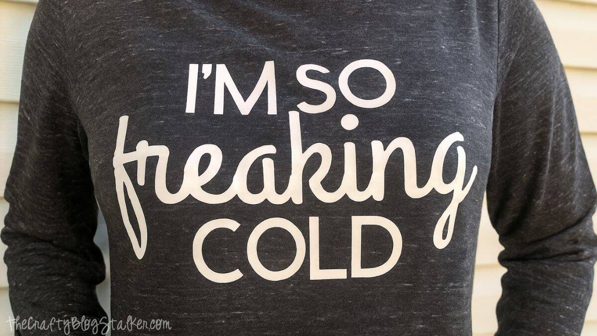 A close-up of an individual donning a long-sleeve sweatshirt with a witty winter phrase printed on the front, captured outdoors against a house backdrop.