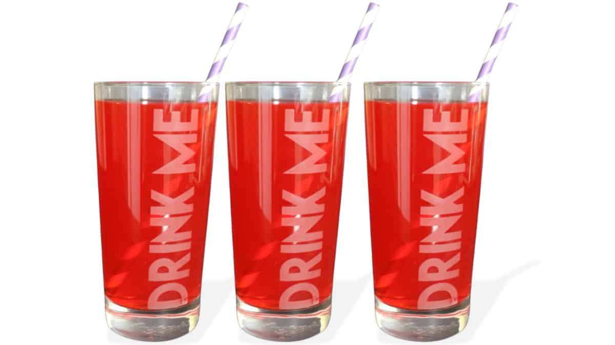 Three tall etched drinking glasses saying 'Drink Me,' each filled with a beverage and paper straw, displayed side by side.